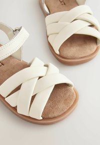 Vita lädersandaler med ett korslagt mönster, en mjuk fotsula av brunt nubuck och en rund gummisula. Justerbart rem med metallbeslag.