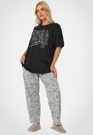 Femme portant un haut noir oversize avec un imprimé sur le thème du petit-déjeuner à Paris et un pantalon gris clair avec des motifs assortis de nourriture et de la tour Eiffel, plus des chaussons gris.