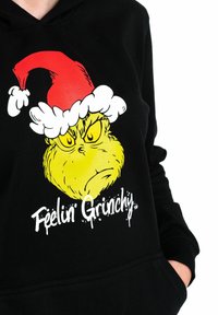 Felpa nera con cappuccio che presenta un graffito del Grinch con un cappello da Babbo Natale rosso. Il design include accenti gialli e bianchi con il testo "Mi sento Grinchy."