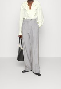 Person klädd i högmidjade grå vida byxor, krämfärgad blus med volanger, svarta fyrkantiga skor och hållande en svart bucket-väska.