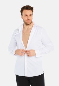 White button-up shirt made of cotton, featuring a spread collar, long sleeves, and a slim fit, worn over a beige undershirt.