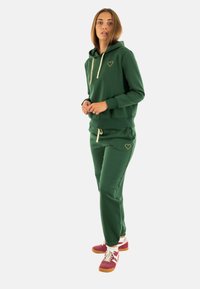 Grøn bomulds hoodie med et guld hjerte-logo og snøre, parret med matchende grønne joggers med et broderet hjerte. Røde og hvide sneakers.