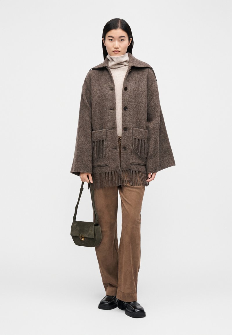 Brun uldfrakke med frynser, store forlommer og knaplukning. Bæres over en beige rullekrave og kombineret med brune bukser og en grøn taske.