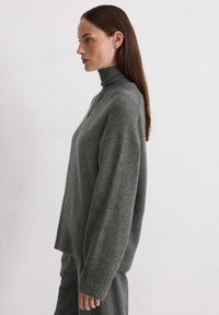 Szary, oversize'owy sweter dzianinowy z golfem, z prążkowanymi mankietami i dołem, pokazany z boku na jednolitym tle.