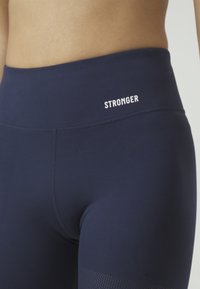 Marina yogabyxor med bred midja. Ordet "STARKARE" är tryckt i vitt på midjan. Har en texturerad detalj nära fållen.