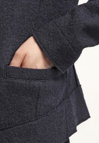 Détail d'une veste en feutre bleu marine avec une main placée dans une poche latérale, présentant un tissu texturé et une coupe superposée asymétrique.