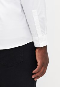 Camicia bianca con bottoni, abbinata a pantaloni neri. Presenta una texture di tessuto liscia e due bottoni sui polsini.