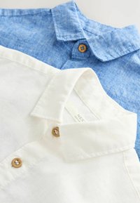 Sono esposte due camicie colletto. La camicia superiore è azzurro chiaro, mentre quella inferiore è bianca, entrambe con bottoni in legno e una trama in lino.