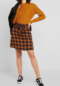 Pull orange en maille avec motif texturé, associé à une jupe à carreaux bleu et orange avec un lien à la taille. Des baskets noires complètent la tenue.