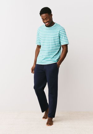 Smiling barefoot man wearing a light blue striped t-shirt and dark blue pants standing on a light carpet against a plain wall.
