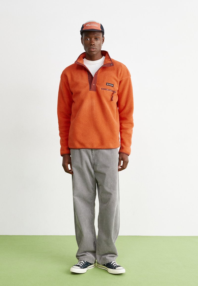 Pullover arancione in pile con colletto marrone e tasca con zip, abbinato a pantaloni a gamba larga grigi a righe e sneakers nere.