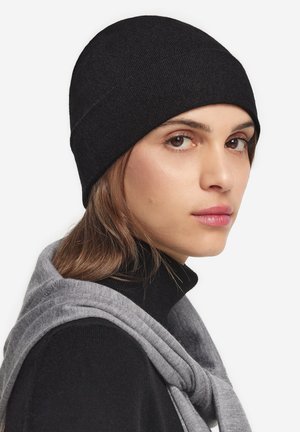 Beanie nera in maglia dal texture liscia, design aderente e corona arrotondata. Abbinato a una leggera sciarpa grigia appoggiata sulle spalle.