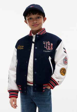 Dreng iført navy- og hvid varsityjakke med røde stribede manchetter og mærker, hvid sweater, blå kasket og blå jeans.