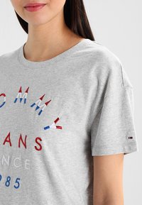 Kobieta ubrana w jasnoszary t-shirt Tommy Jeans z haftowanym czerwono-biało-niebieskim napisem i małym logo flagi na rękawie.