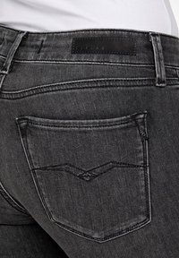 Jeans de mezclilla negra con una textura suave, que presentan un parche de logo en la cintura y un único bolsillo trasero con costuras en contraste.