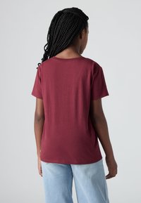 Cilvēks ar garām bārdām, ģērbies vienkāršā maroon krāsas īsrokainā krekliņā un gaiši zilās džinsās, skatās prom pret vienkāršu fona.