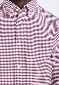 Camicia a maniche lunghe a quadri rossi e bianchi con colletto classico. Presenta bottoni bianchi e un piccolo dettaglio con logo nell'angolo in alto a sinistra.