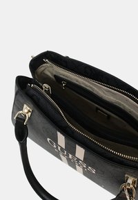 Sac à main noir avec une fermeture éclair argentée, dévoilant plusieurs compartiments et le logo "GUESS" sur le côté avec des rayures blanches.