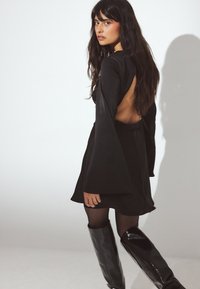 Robe noire mini avec de longues manches fluides et un dos ouvert, associée à des bottes noires brillantes montantes. Texture lisse, détails minimalistes.