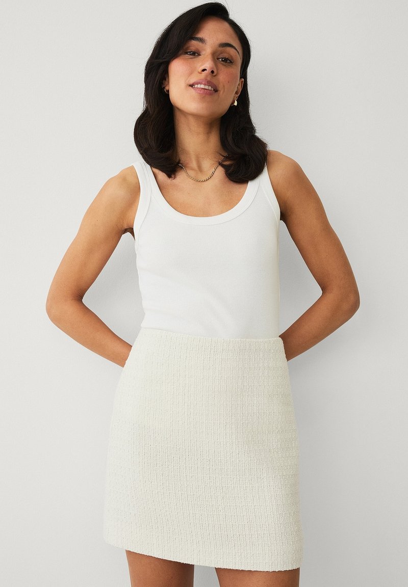 Vrouw met donker haar die een wit mouwloos geribd topje en een getextureerde off-white minirok draagt, staand met haar handen achter haar rug.