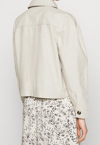 Jasno beżowa krótka kurtka z kołnierzykiem koszulowym, zapięcie na guziki i prosty krój. Gładki materiał z minimalnymi detalami szwów.