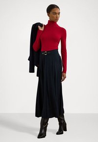 Rdeča majica z dolgimi rokavi in polo ovratnikom, kombinirana s temno modro maxi krilo z pasom in zlatim sponko. Model nosi črne gležnjarje.