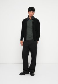 Μαύρο ζιβάγκο με φερμουάρ και ψηλό γιακά, πάνω από ένα σκούρο πράσινο πουκάμισο; συνδυασμένο με φαρδιά μαύρα παν trousers και μαύρα παπούτσια.