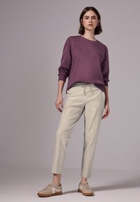 Paarse sweater met lange mouwen en ribbels aan de manchetten, gecombineerd met lichtbeige tapered broeken met een trekkoord in de taille, gedragen met beige sneakers.