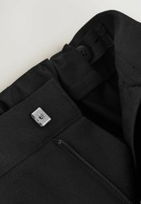 Schwarze Hose mit einem Reißverschluss und einem metallischen Ziehband sowie einem Knopfverschluss. Hergestellt aus strukturiertem Stoff mit einem glatten Innenfutter.