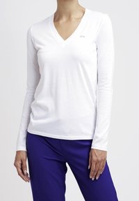 Weißes Langarm-V-Ausschnitt-Shirt aus weichem Stoff, mit einem kleinen Logoakzent auf der Brust, kombiniert mit blauen Hosen.