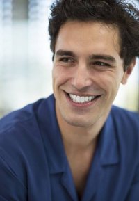 Uomo sorridente in una camicia blu con capelli scuri, corti e ricci, con un sorriso luminoso e pelle chiara, su uno sfondo leggermente sfocato.