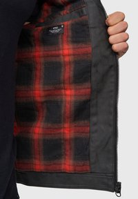 Chaqueta negra con cremallera que presenta un forro a cuadros en rojo y negro, textura exterior suave y un bolsillo interior con etiqueta.