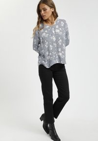 Blusa floreale grigio chiaro con motivi bianchi, vestibilità rilassata, scollatura a V, abbinata a jeans neri e stivali alla caviglia. Tessuto liscio, design casual.