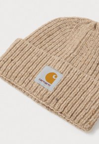 Gorro de punto acanalado Carhartt en color beige con puño doblado y parche cuadrado con logo en amarillo y blanco sobre un fondo blanco.