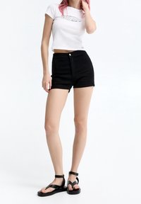 Camiseta blanca recortada con texto impreso, pantalones cortos de mezclilla negros y sandalias negras con tiras y suela gruesa; textura suave en general.