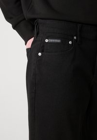 Jean en denim noir avec une texture lisse, dotés de quincaillerie en argent, d'un patch logo Calvin Klein et d'un design classique à cinq poches.