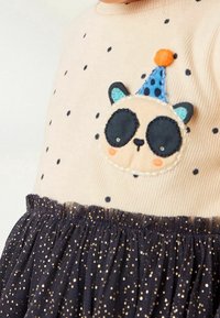 Un haut cr�émeux avec des pois noirs, mettant en avant une appliqué panda portant un chapeau de fête bleu. Jupe en tulle noir avec des paillettes dorées au bord.