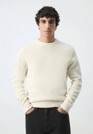 Joven con cabello rizado y oscuro, vistiendo un suéter de punto color crema y pantalones negros, de pie con las manos en los bolsillos frente a un fondo liso.