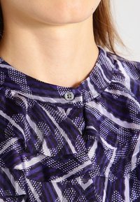 Detailní záběr na krk osoby, oblečené v košili s fialovým vzorem a knoflíkem na límci, zdůrazňující texturu a design látky.