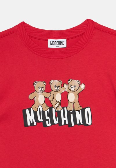 Κόκκινο παιδικό πουκάμισο με τρία παιχνιδιάρικα αρκουδάκια πάνω από έντονα λευκά γράμματα που σχηματίζουν τη λέξη "MOSCHINO" σε μαύρο φόντο.