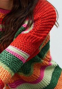 Gebreide trui met levendige strepen in rood, groen, roze en oranje, met een gestructureerd patroon en een brede, geribde halslijn.