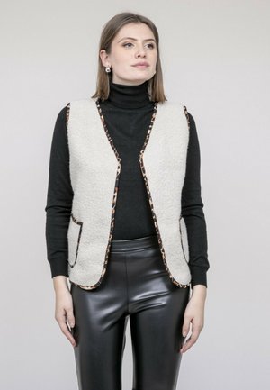Femme portant un gilet blanc texturé avec bordure léopard, un pull à col roulé noir, un pantalon en simili cuir noir et des boucles d'oreilles en perles, debout devant un fond gris.