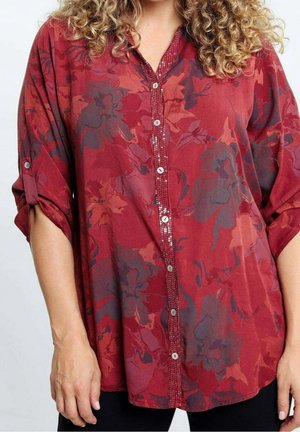 Mujer con camisa suelta roja de botones y estampado floral, mangas remangadas y cabello rizado visible en el borde superior.