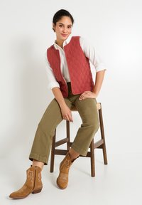 Gilet matelassé rouge avec un col rond, associé à une chemise blanche à boutons et un pantalon large vert olive. Portant des bottes de cowboy beiges avec broderies.