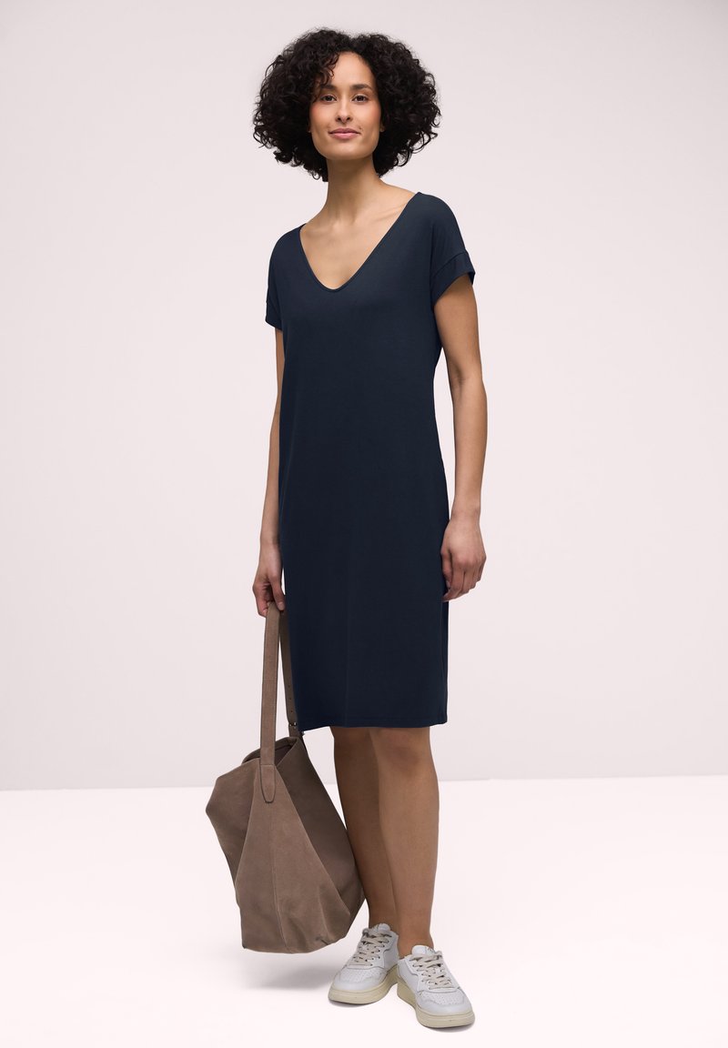 Femme aux cheveux bouclés portant une robe bleu marine à manches courtes, des baskets blanches, et tenant un grand sac fourre-tout marron, debout sur un fond uni.