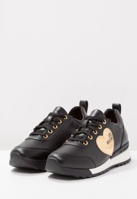 Black leather sneakers featuring gold eyelets, black laces, a beige heart accent with "LOVE MOSCHINO," and a contrasting white sole.