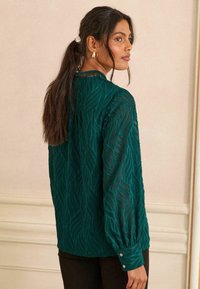 Blusa teal con texture a onde, maniche lunghe, polsini con bottoni e colletto alto. Abbinata a pantaloni neri e indossata con i capelli raccolti.
