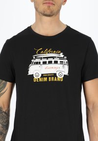 Musta puuvillaita paita, jossa on valkoinen ja keltainen grafiikka vintage-vanmista, jossa lukee "California dreaming" ja "DENIM BRAND."