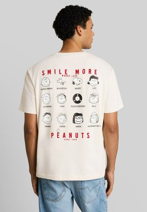 Mann trägt ein weißes Hemd mit Peanuts-Figuren und Text "Smile More Worry Less" und "Peanuts Since 1950" auf dem Rücken.