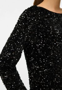 Svart långärmad topp täckt med små, glänsande svarta paljetter; har en rundad halsringning och en texturerad yta.